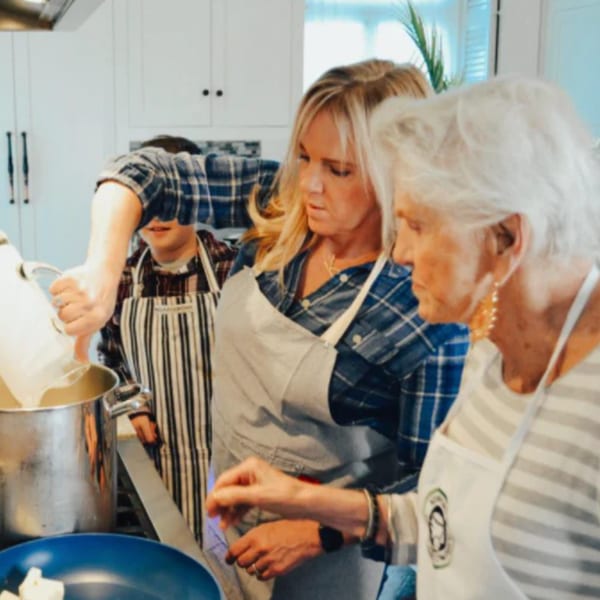 Three people in aprons cooking together in a bright kitchen, pouring ingredients into a pot.