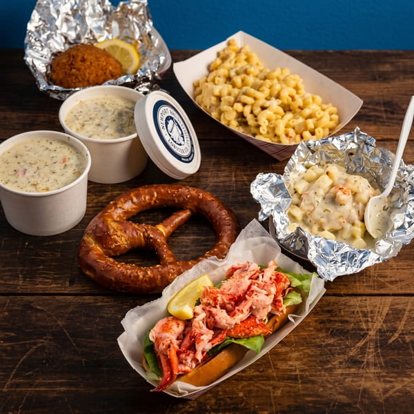 A lobster roll, pretzel, mac and cheese, chowder, and lobster bisque on a wooden table.