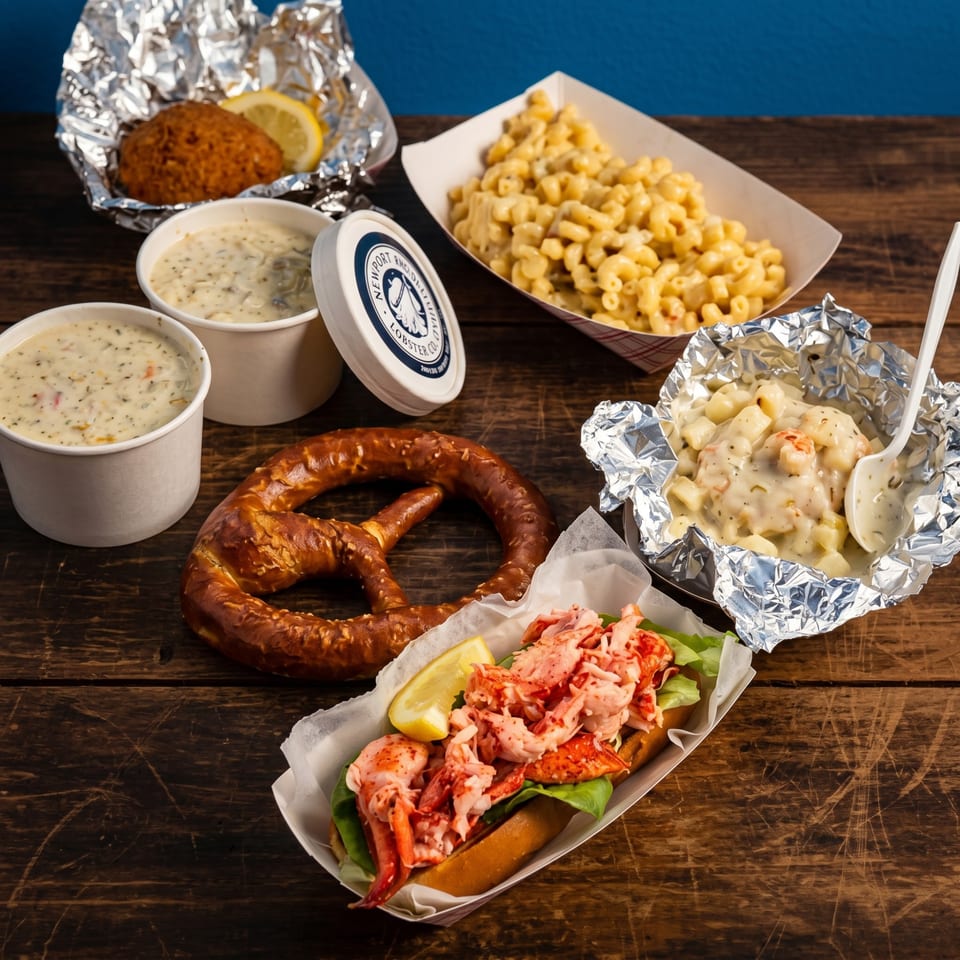 A lobster roll, pretzel, mac and cheese, chowder, and lobster bisque on a wooden table.