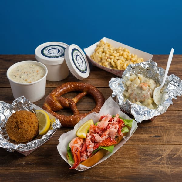 A lobster roll, chowder, mac and cheese, a pretzel, and a fried crab cake on a wooden table.