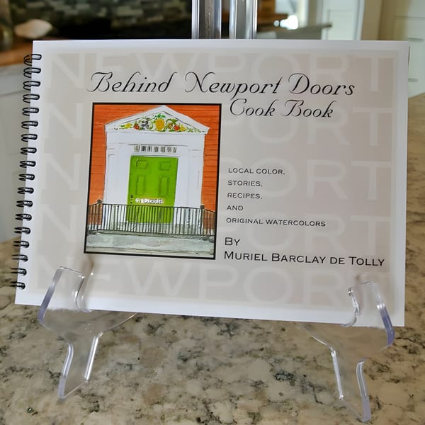 A spiral-bound cookbook titled "Behind Newport Doors Cook Book" on a clear stand.